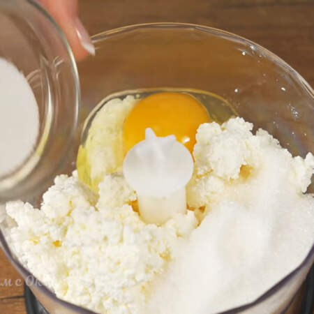 В измельчитель кладем половину творога, так как за один раз все не помещается. Разбиваем 1 яйцо, насыпаем половину сахара и добавляем ванильный сахар.