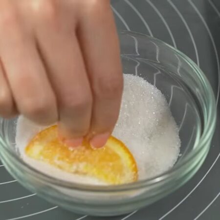 Берем примерно 100 г сахара и обмакиваем в нем подготовленные апельсиновые дольки.