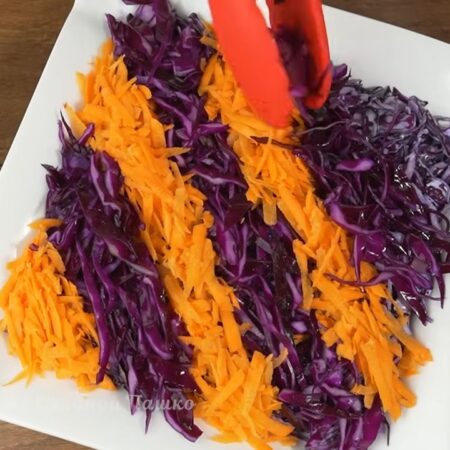 Украшаем салат. По диагонали на салат выкладываем полоску отложенной краснокочанной капусты. Затем сбоку выкладываем тертую морковь. Таким методом чередуя полосы украшаем весь салат.