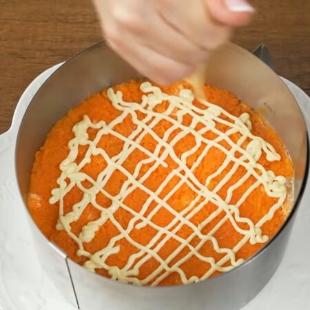 Следующим слоем кладем тертую морковь. Равномерно ее распределяем и утрамбовываем ложкой. Немного солим. Наносим сеточку из майонеза.
